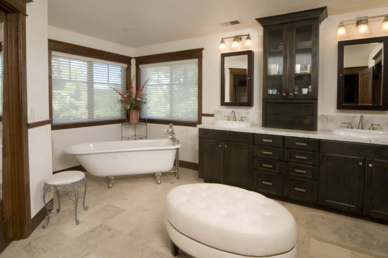 acrylic clawfoot bathtub with floor-mounted tub filler and large oval tufted ottoman with nailhead trim and dark walnut wooden cabinet with reeded glass insert and mosaic edge countertop with two overmount sinks