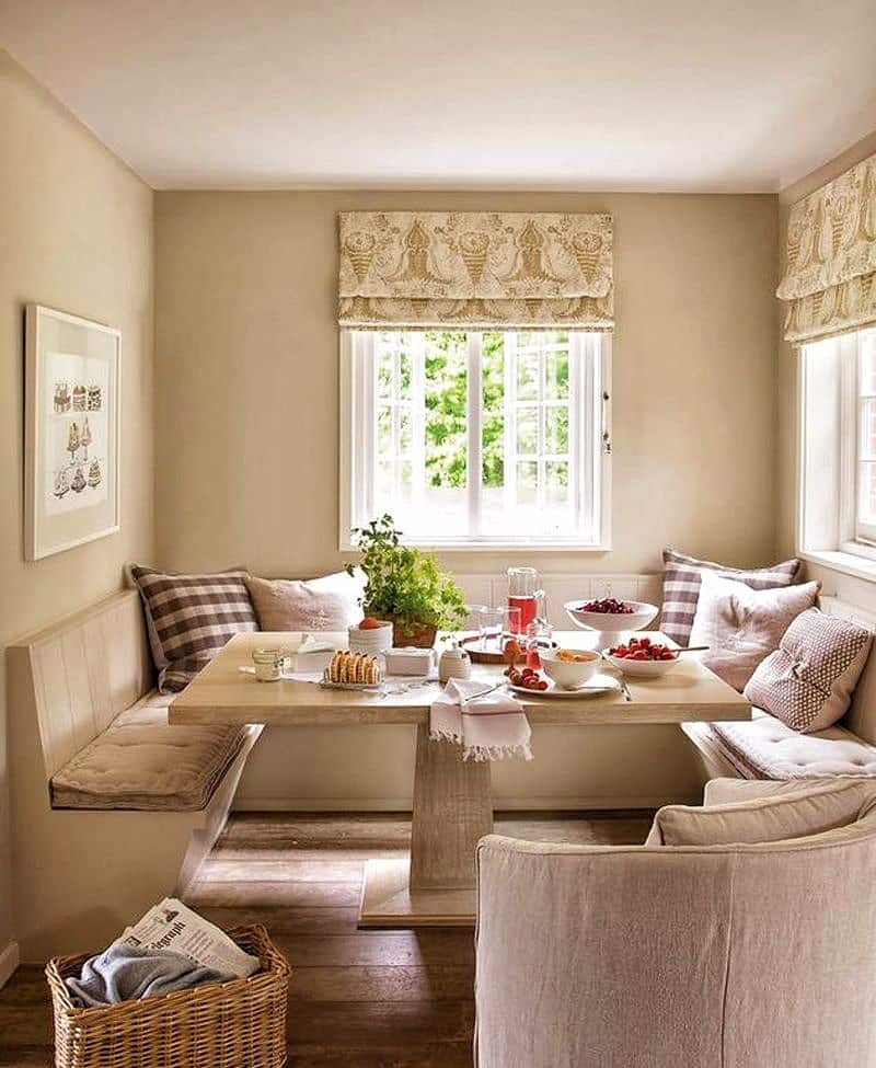 beige breakfast nook benches with tufted cushions and rectangular single pedestal quartzite table with weaved wicker basket and upholstered barrel chair with matching pillow with classic printed fabric Roman shades