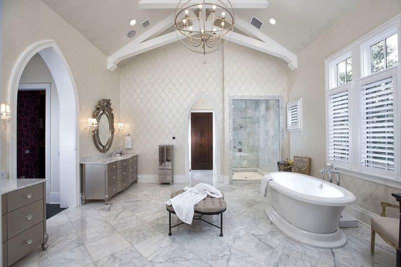 cathedral ceiling with decorative beams and metal orb chandelier and gray cabinets with crystal glass knobs and white window shutters with oversized tufted beige stool with black metal legs