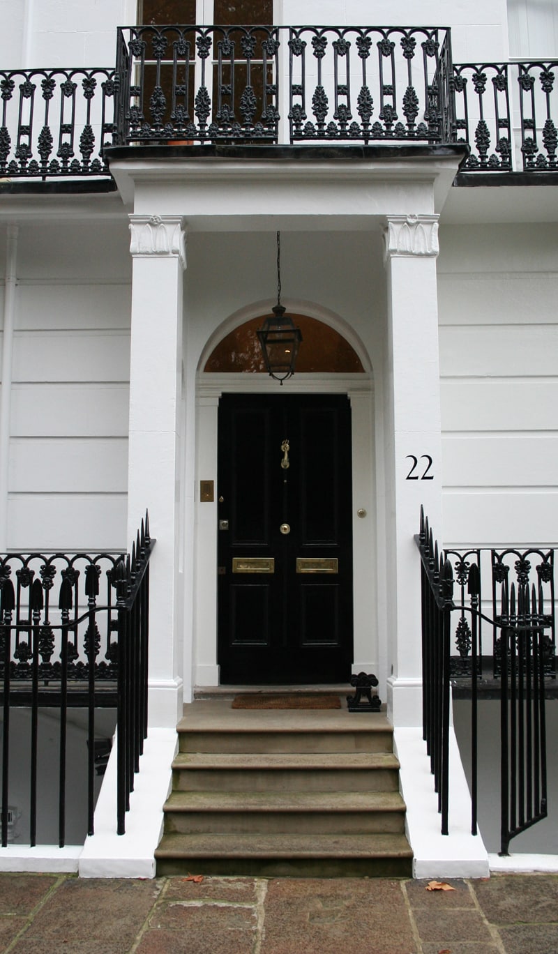 double brass mail slots on a recessed panel door and balcony over entry with house marker on portico column and black cast iron railings with spear points by the steps