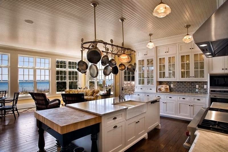 glass muntin cabinets with all-wood upper cabinets and smoke color brick backsplash with ocean view windows with transom and white marble island with apron-front sink and hanging brass pot rack