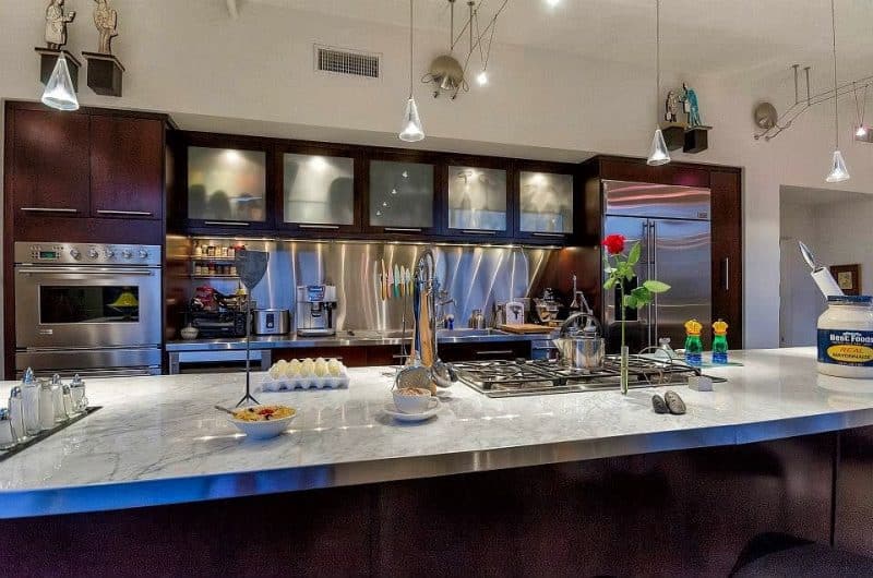 lift-up frosted cabinet doors with interior lighting and illuminated stainless steel backsplash with white quartzite island overhang with range cooktop and decorative wood sculptures