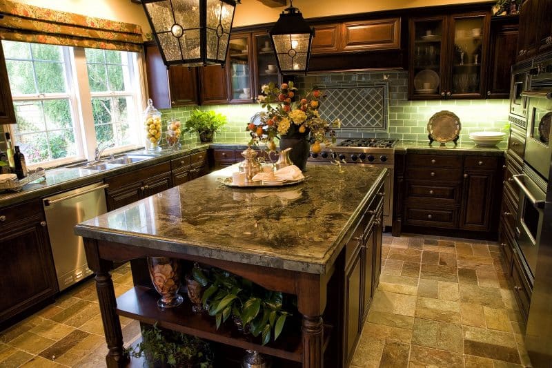 seeded glass cabinet inserts and chiseled edge travertine finish flooring with marble counter and island tops and mint subway tile backsplash and single hung windows with white muntins and twin pendant lights with same seeded glass as the cabinet inserts