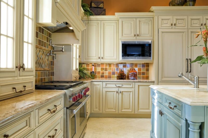 tiled backsplash in various earth tone hues with creamy white recessed-panel cabinetry and oil rubbed bronze handles with terra cotta walls above cabinet crown molding