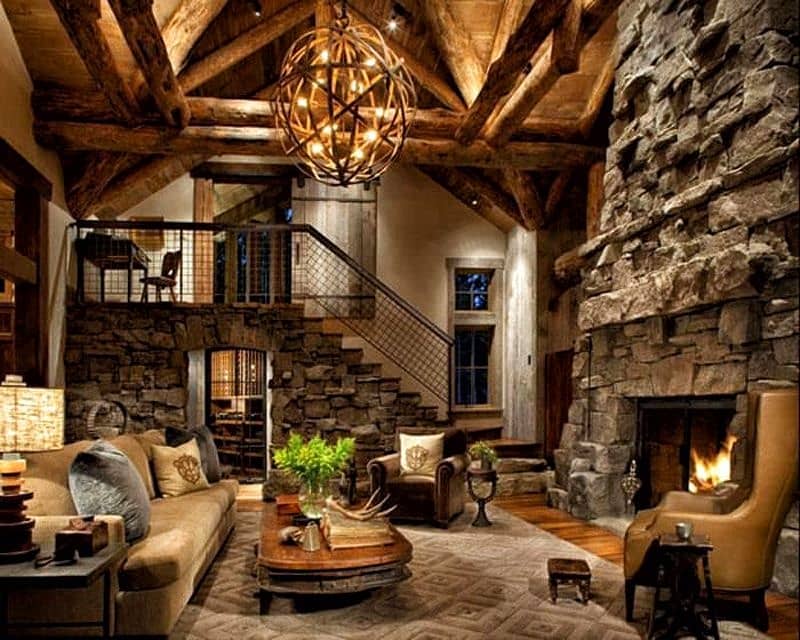 vaulted ceiling with timber beams and trusses with floor-to-ceiling stone fireplace and leather wingback chair with vintage foot stool with barn door for upstairs bedroom and large orb chandelier