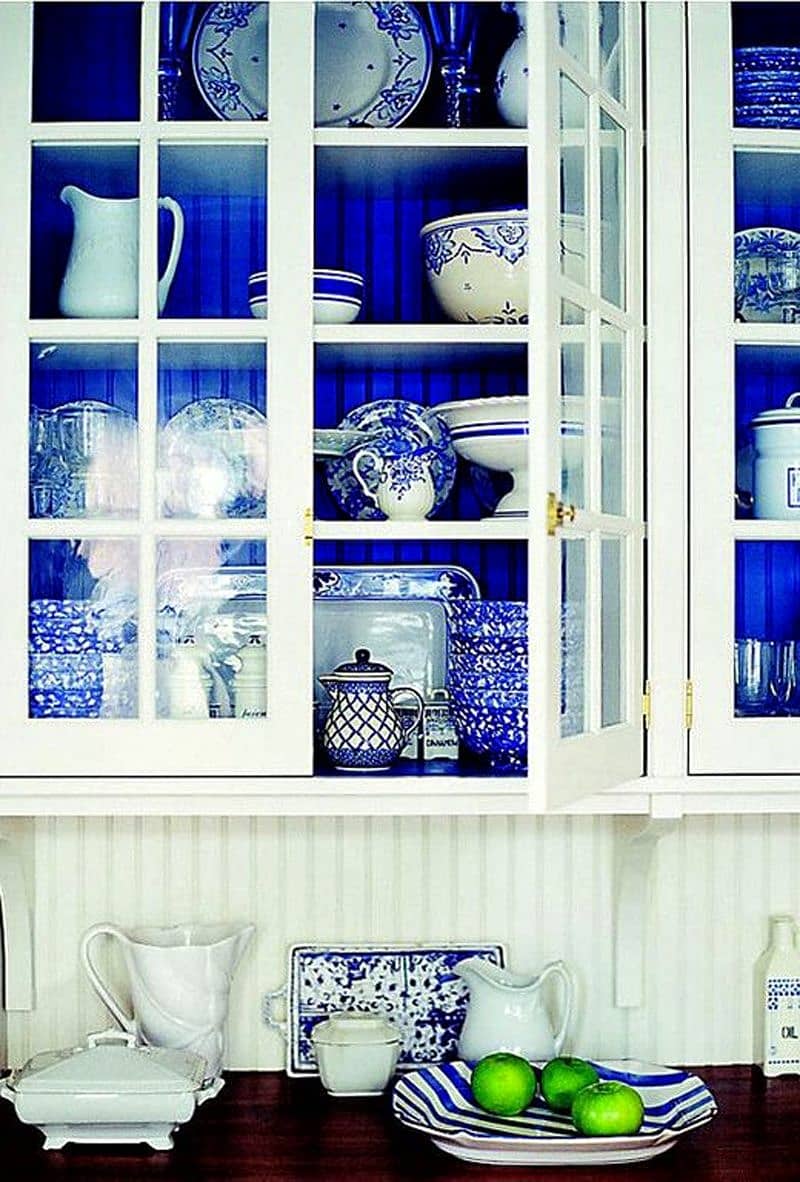 white glass cabinets with painted blue shelf and back panel with blue and white porcelain and dark solid wood countertop with beadboard wooden wall and thin brass cabinet hardware
