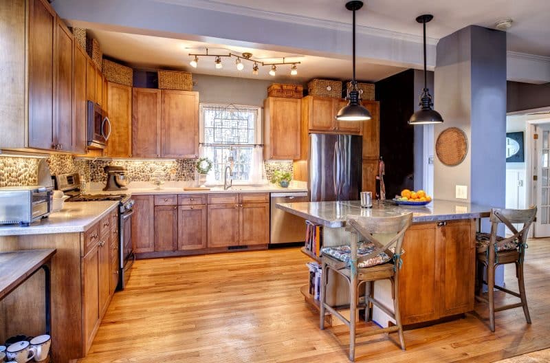 granite topped island with curved open shelving and X-back wooden counter chairs with tie back seat cushions and pebble backsplash with wicker storage baskets above upper cabinets and lipped round wicker tray on column