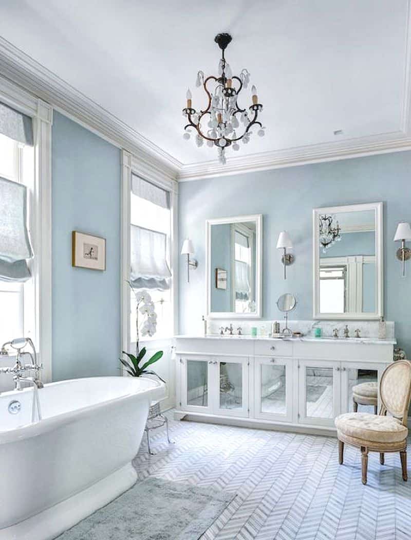 baby blue walls with white marble countertop with sidesplash and sheer baby blue fabric Roman shades with mirrored white base cabinets and cream round armless chair with reeded legs and gray and white chevron tiled floor