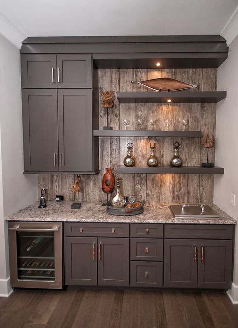 beige granite countertop with built-in ice bucket and rustic wood plank backsplash with matte gray with brownish hues cabinets and amber handles and square knobs on base cabinets with under-counter wine cooler