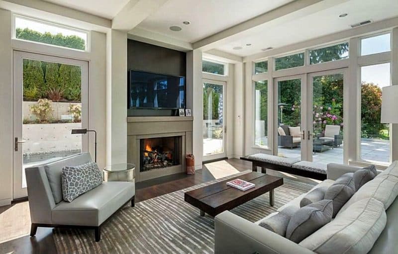 light gray lounge sofa with matching throw pillow and bench cushions and open fireplace with spark guard curtains and dark gray TV niche above fireplace with glass walls with garden view