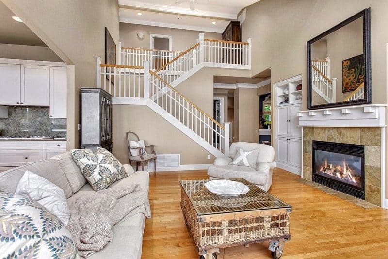 matching white modern shaker cabinets in kitchen and beside fireplace with steel gray granite slab backsplash and cane wicker glass topped coffee table on casters and oversized black framed mirror above clearview fireplace