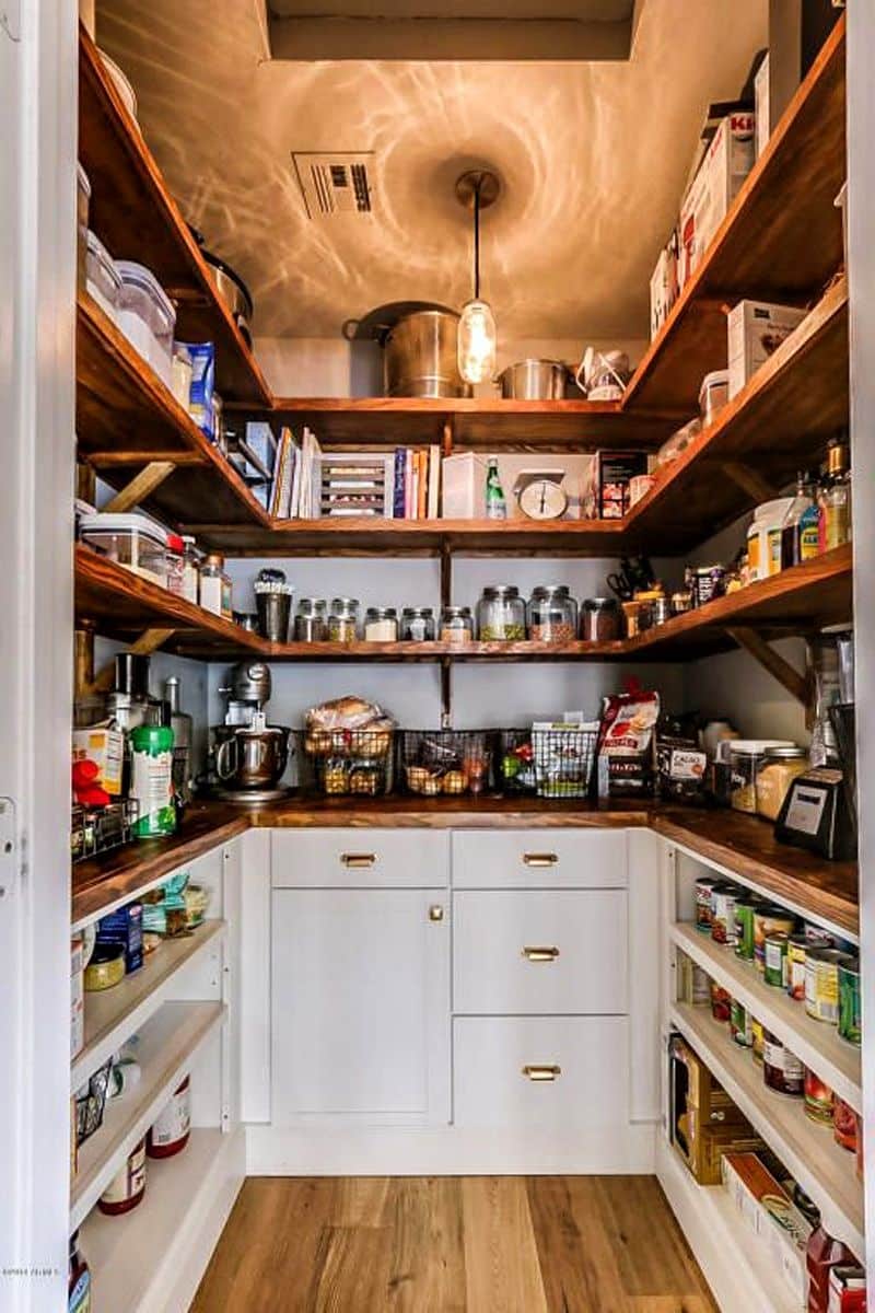U-shaped walk-in pantry with dark-stained hickory shelves and white lower cabinet and drawers with awning cup pulls in gold finish with DIY glass bottle pendant light and multi-sized mason jars