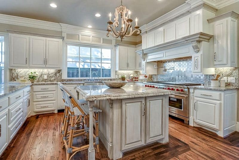 white upper cabinets with glazed crown molding and under-cabinet lighting with glacier white granite countertops and backsplash with French cafe counter chairs and wood floor inlay following island shape