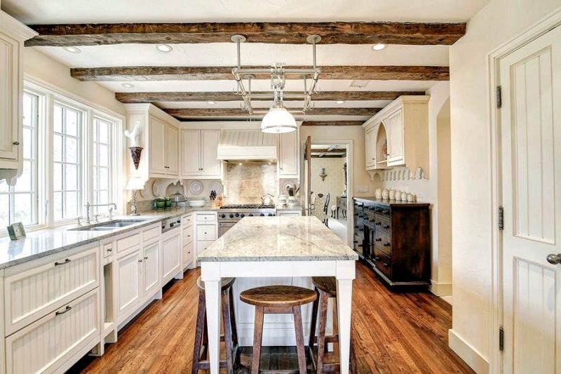 antique red oak floors and antique apothecary sideboard with drawer knobs and beadboard panels on door cabinets and range hood with reclaimed wooden beams on ceiling and wooden stools tucked under kitchen island