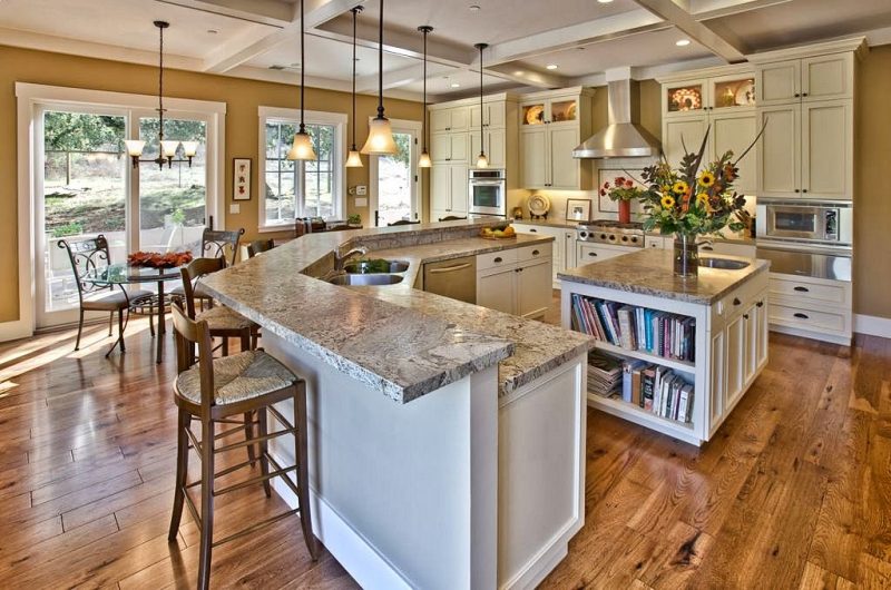 ash hardwood floors and white coffered ceiling with recessed lights and V-shaped two-tiered island granite countertop with built-in bookshelves in square kitchen island and six brown counter rush seats