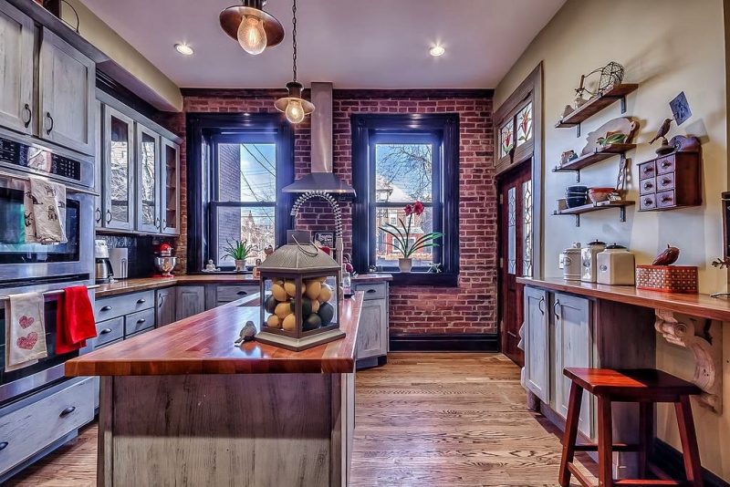 ash hardwood floors with two single-hung windows on exposed brick wall and light gray milk painted cabinets with wooden island countertop with glass lantern fruit holder and stained glass transom window