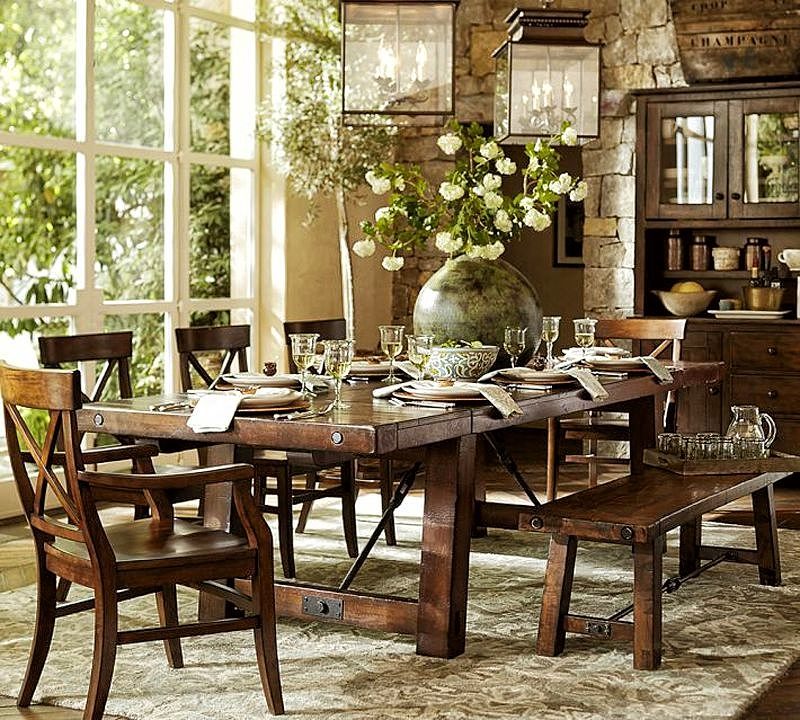 dark walnut dining table with wrought iron brackets and oversized round table vase with white carnations and square glass pendant lantern with weathered brass frame and walnut cupboard against stacked stone wall