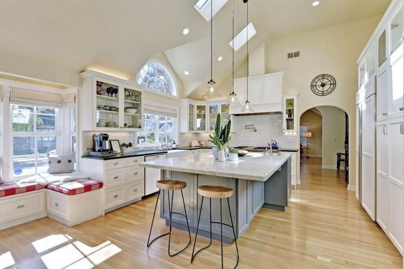 light maple hardwood floors with L-shaped window seat with three-toned red plaid cushions and skeletal Roman numeral clock above archway to living room with two small square skylights and marble top island with overhang