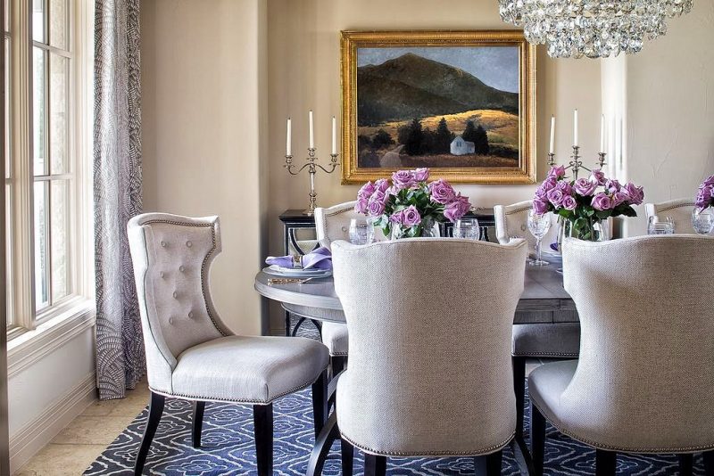 tufted wingback dining chairs with nailhead trim and antique gold-framed painting above mirrored sideboard with lavender roses in glass pedestal vases and crystal chandelier above oval wooden dining table