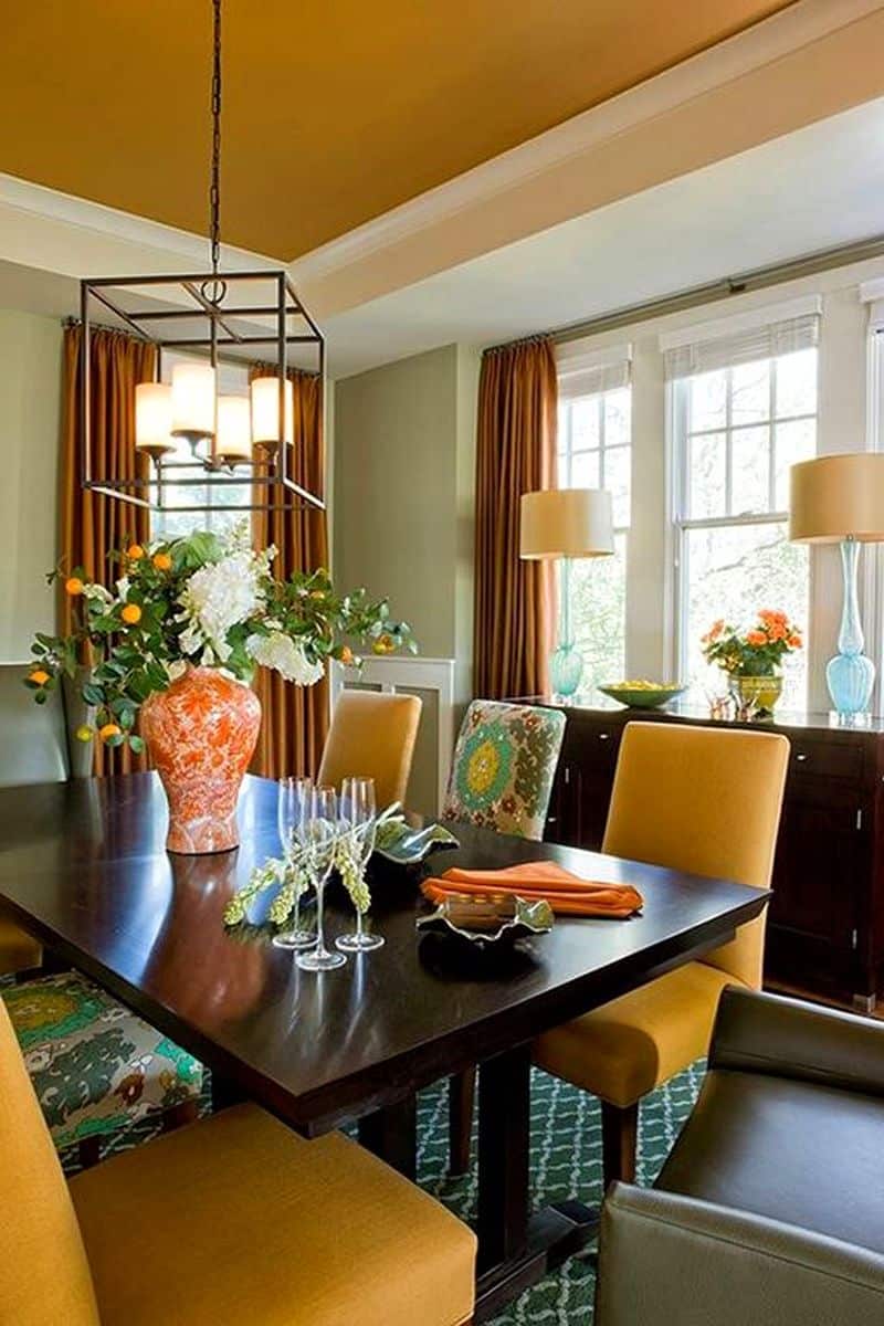 3 mustard yellow dining chairs matching the ceiling and pale sage green walls with beige lamp shades with reeded glass base and hand painted orange and white porcelain vase with green trellis area rug