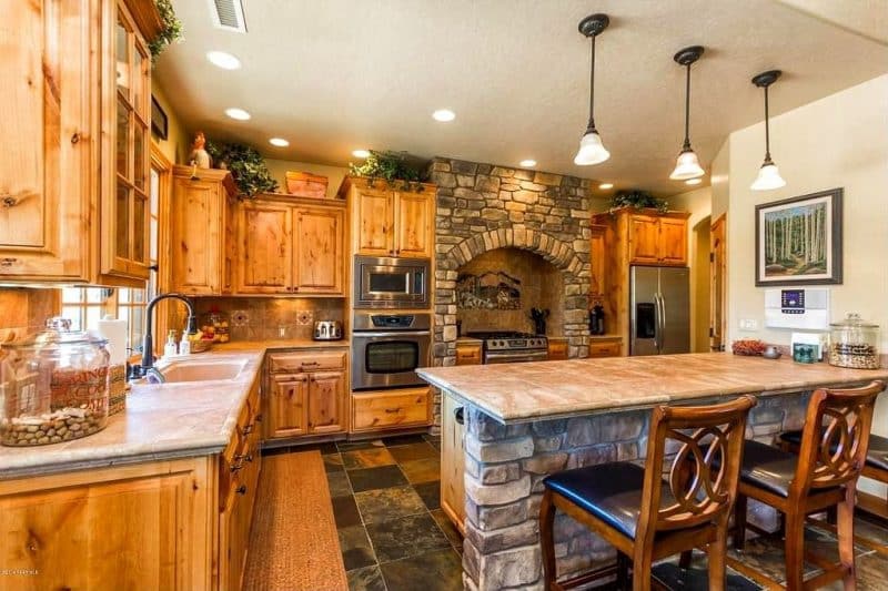 pine wood cabinetry and honed travertine tile island countertop with white washed stacked stone base and stacked stone stove alcove with hand painted tile mural backsplash and sisal floor runner on slate tiled floor