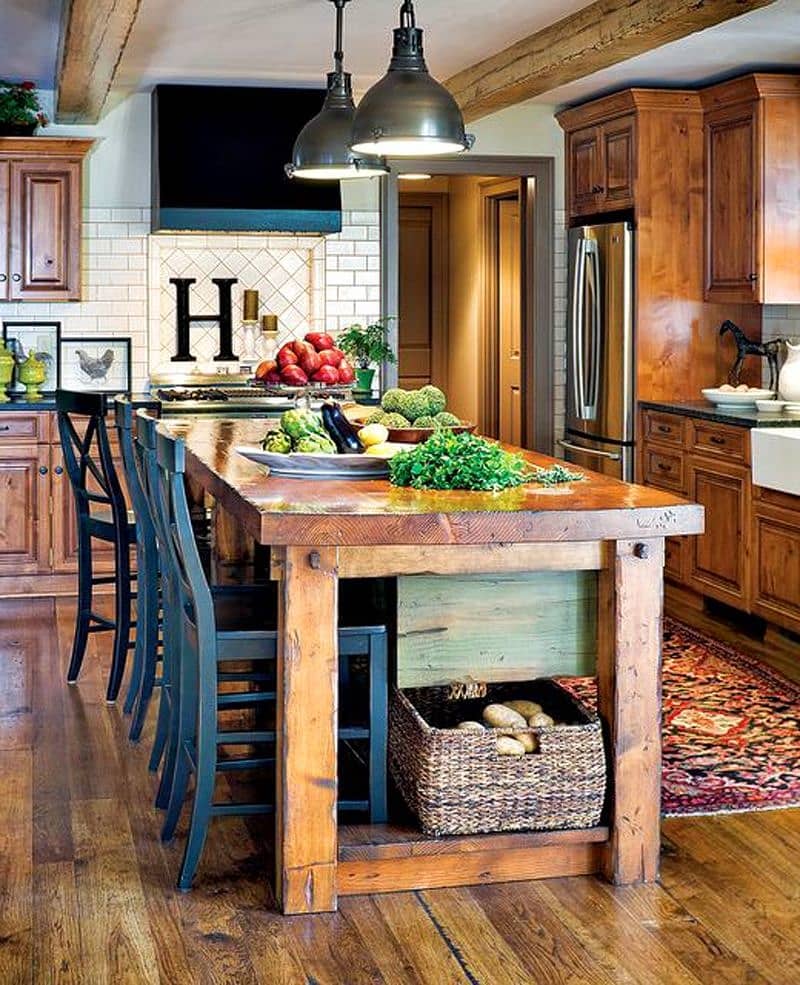 varnished wooden island table with Mediterranean blue cross back chairs and recessed stove niche with white porcelain tiles in diamond pattern and wicker storage basket under island countertop with reclaimed timber on white ceiling