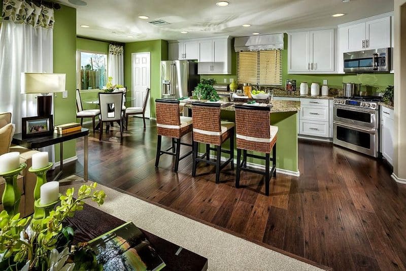 4 white upholstered dining chairs with cut-out backrest and 3 wicker counter chairs with white tie-back cushions and Crystal Gold granite countertops with white curtain with attached valance in dining area window matching the fabric Roman shades above kitchen sink and walls and island base in Benjamin Moore’s ‘Napa Vineyard’ green and handscraped engineered hardwood floor