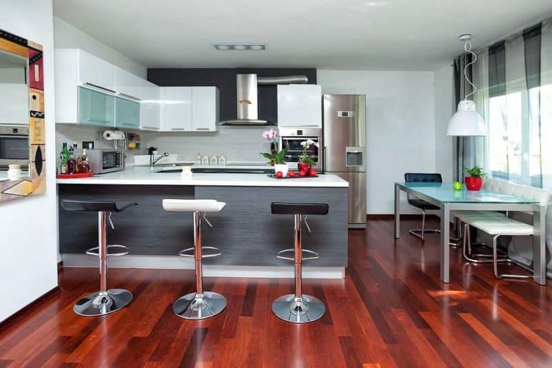 U-shaped kitchen with white Corian countertop and corner sink with shelf with black and white low back swivel bar stools with chrome pedestal base and back-painted glass topped dining table with black and white tufted leather dining chairs with L-shaped legs and wall-mounted mirror with hand painted geometric design on wood frame with Brazilian cherry hardwood flooring