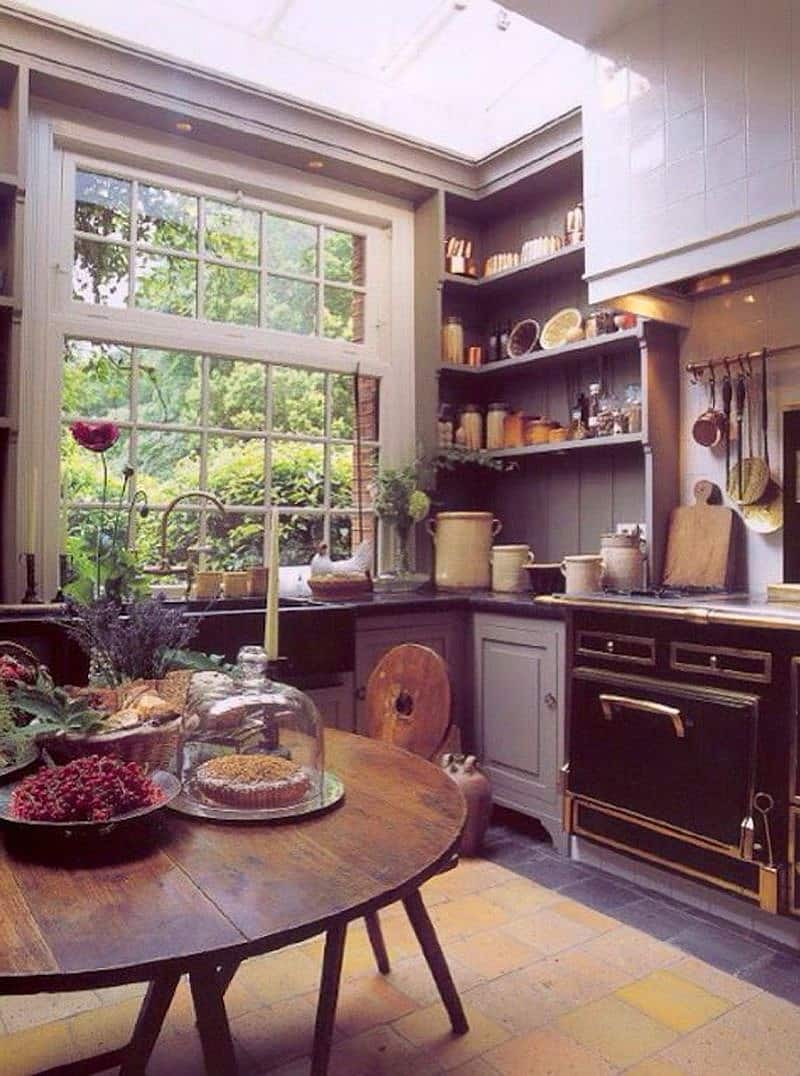 antique round back dining chair with small circular cut-out and metal cake tray with glass cloche on round antique wooden dining table with assorted glazed ceramic jars on soapstone countertop and wooden open shelves and white porcelain tiled range hood with large skylight providing natural light to kitchen and square brick patterned travertine floor with soapstone tile border