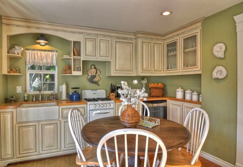 chocolate glazed raised panel cream cabinets with rope molding and butcher block countertops with wicker storage baskets and woven bamboo vase with silver dollar bouquet on round wood dining table and open shelves with arch valance over farmhouse sink with resin mermaid on clam shell throne wall decor above white stove and 2 resin ammonite fossil seashells on olive green wall