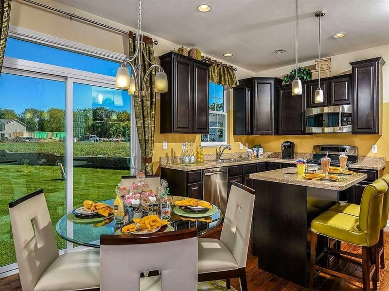 dark wooden cabinets with built-in stainless steel kitchen appliances and small kitchen island with 2 slim lime green leather slipcovered counter chairs and vintage storage boxes above upper cabinet with dining table topped with round beveled glass and surrounded by chairs with cut-out backrest and glass sliding doors to patio with full view transom