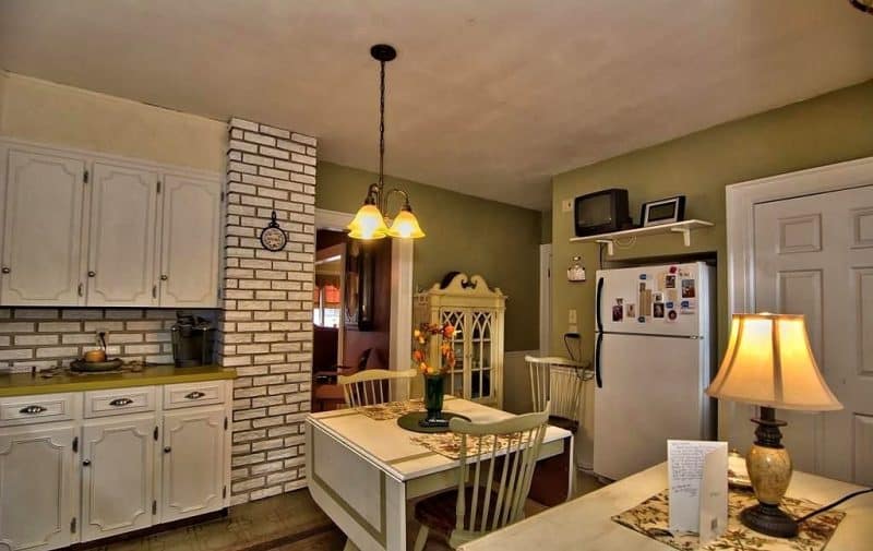 glazed faux brick wall and backsplash with contrasting dark grout and olive green walls with white doors calling back drop-leaf dining table with olive green laminate countertop with exposed hinged white cabinets and white TV shelf above refrigerator alcove and small cream display cabinet with scrolled pediment and 11-lite Gothic mullion cabinet doors