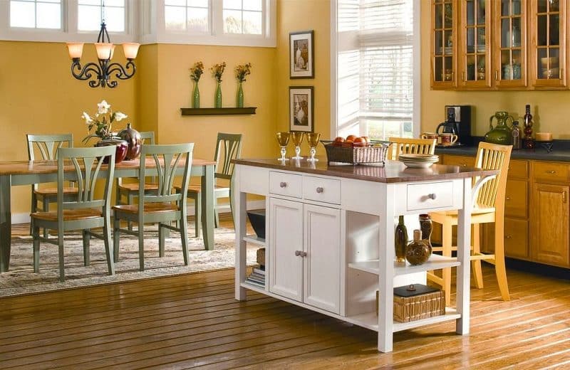 pale sage dining chairs and satin finish wood topped island with white drawers, cabinets, and built-in open shelves with soapstone countertop with sidesplash and 3 slim olive green bottle-shaped vases with Butter Daisies on slim floating shelf and 3 amber glass goblets with clear glass base calling back glass bottles and jars on island shelf