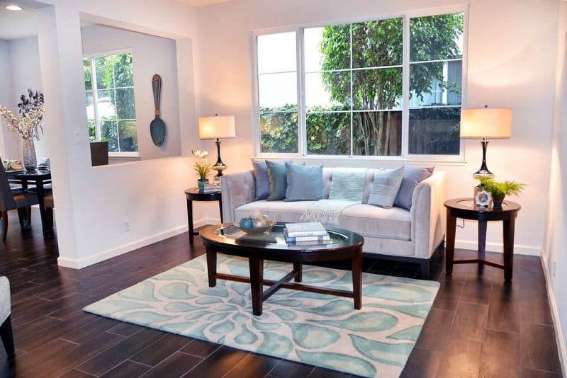 sea salt tuxedo sofa multi-toned blue throw pillows and throw with living room-kitchen pass through window showing the large decorative peacock blue milk painted spoon on kitchen wall and white and shades of aqua teardrop patterned area rug calling back throw rug and painted decorative wooden balls in hammered aluminum bowl with triangular base and 2 genie bottle table lamps with black and gold ceramic painted base on end tables matching the round glass topped wooden coffee table