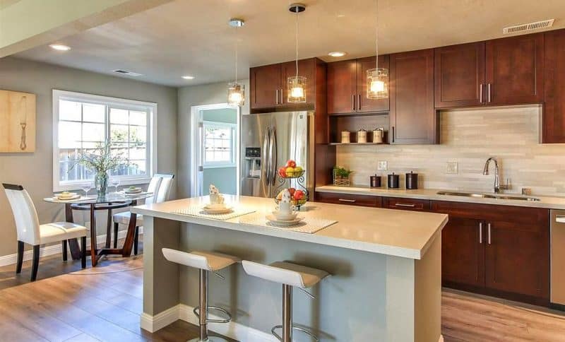white quartz countertop with porcelain linear mosaic tile backsplash and “Bloomfield” dining table with crossing wooden base and beveled glass top with 3-piece oil-rubbed bronze canister set on countertop and 3 glass pendant lights over kitchen island with light gray base and wall-mounted butter knife art on canvas