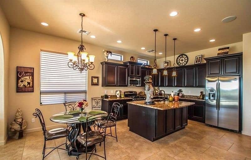 wrought iron dining chairs calling back wrought iron chandelier and 3 pendant lights with coffee brown granite island countertop with curved overhang and skeleton Roman numeral clock above upper cabinet with Italian chef resin statue on kitchen island with cast stone urn with decorative tassel by the archway and 2 clerestory windows above cabinets